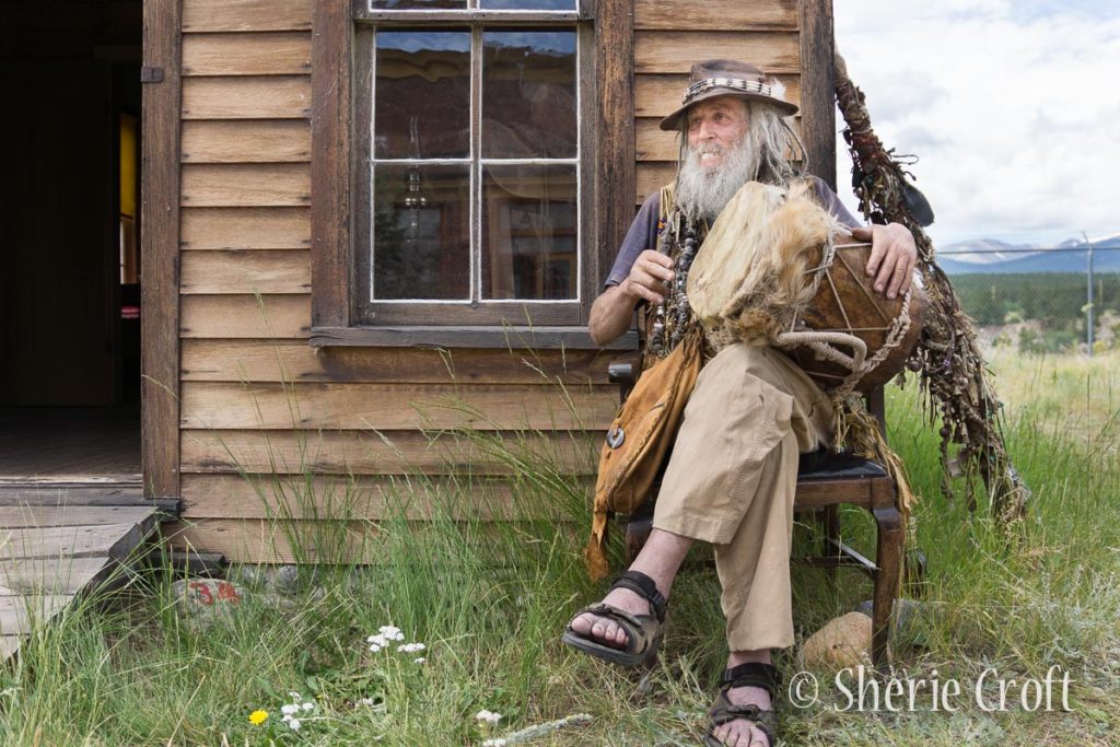 Man with white beard and brown hat seated in front of old weathered building playing a drum.